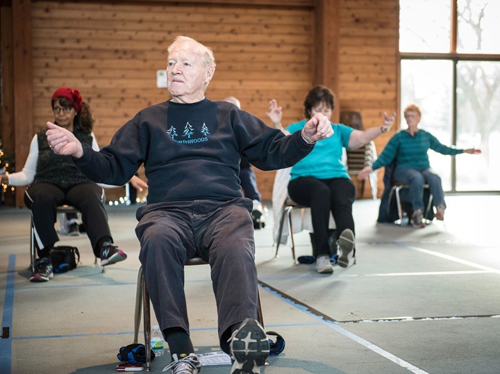 A photo of an exercise class. Four people are seated doing stretches.